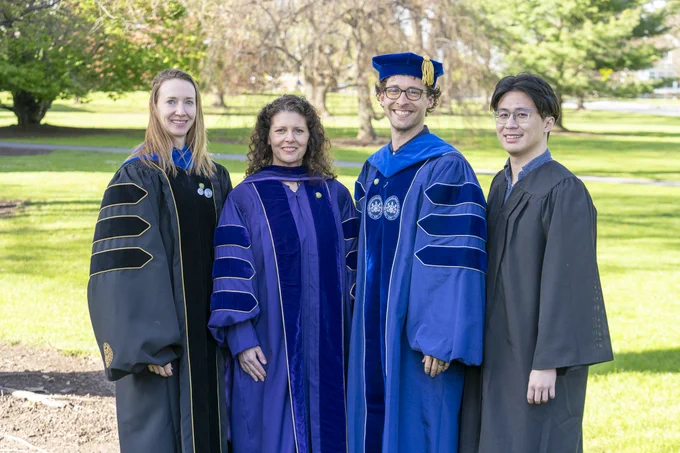 A group of four people dressed in graduation robes stand outdoors on a grassy area, with trees in the background. Two are wearing blue academic regalia with caps, and two are in black robes.