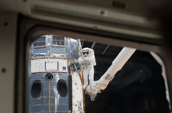 Taken through a window on the space shuttle, the lower portion of Hubble is visible with astronaut Andrew Feustel on the robotic arm completing work.