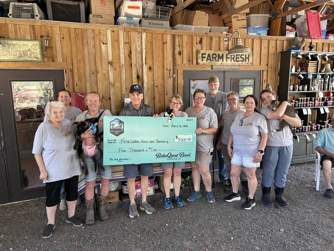 A group of people presenting a $5,000 donation check at a farm. The mood is supportive and cheerful, with everyone in casual attire, and pets are present.