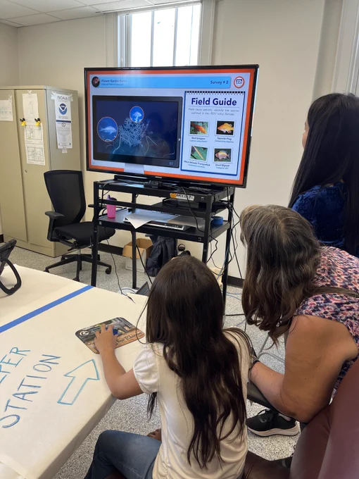 Three people look at a TV screen with photos of underwater creatures and coral on it. 
