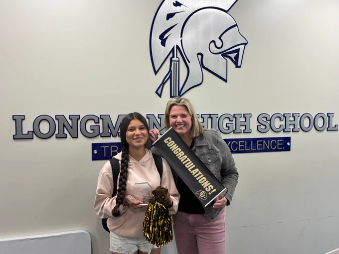 Two people stand together smiling in front of a wall displaying the Longmont High School logo — a Trojan helmet mascot in navy blue and silver — along with the school's motto "Tradition of Excellence."
On the left is a young woman wearing a light pink hoodie and white shorts, with a long braided ponytail and a backpack. She is holding a clear glass award trophy and gold and black pom-poms, along with a small stuffed bear.
On the right is an adult woman with shoulder-length blonde hair, wearing a gray denim jacket and pink pants. She is holding a black banner with gold lettering that reads "Congratulations!" and features the University of Colorado (CU) Buffalo logo.