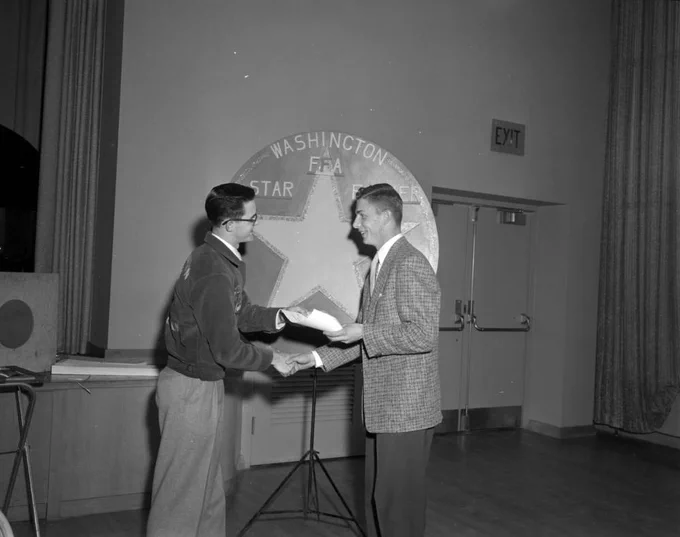 Black and white photo of 2 people shaking hands in front of a sign that says Washington FFA with a large star on it. The person on the left is wearing an FFA jacket and accepts a paper from the other, who is wearing a coat and tie.
