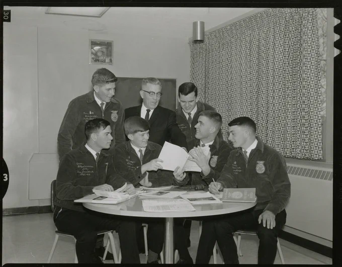 Black and white photo of 7 people sitting or standing around a table looking at papers that two of them are holding. 6 of them are wearing FFA jackets, the 7th is wearing a suit and tie.