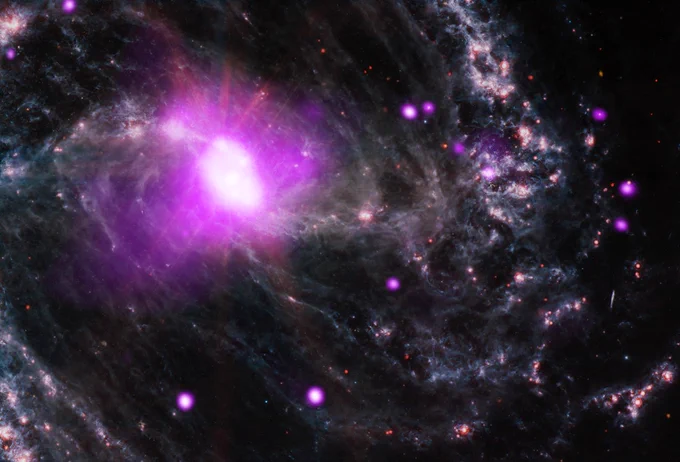 A close up image of spiral galaxy NGC 1365 and the supermassive black hole at its center. Here, the galaxy is shown at a dramatic angle, as if the bright pink core is gazing past our right shoulder. Swirls of pale, grey-blue material, resembling waves in a dark ocean, spiral toward the radiant pink core, which hangs at our upper left. Glowing pink circles, and flecks of red, dot the churning spiral galaxy.