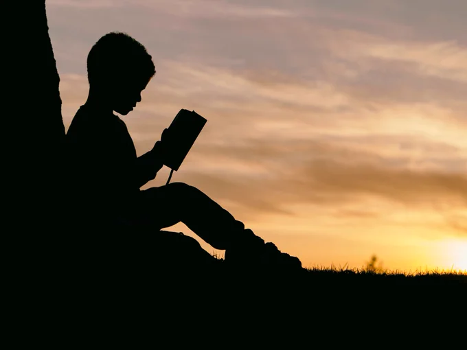Silhouette of a child reading a book in front of a sunrise or sunset