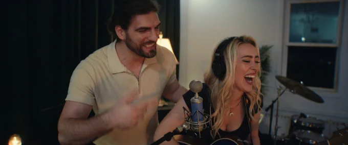 A man smiles behind a laughing woman with headphones, who is singing into a mic. She's playing guitar, with drums visible in the background, in what looks like a home recording studio.