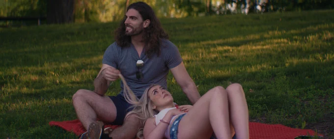 A man sits cross-legged on a blanket in the grass, with a woman lying down, her head in his lap. He's holding her hair. The man has long brown hair and a beard; she has blonde hair.