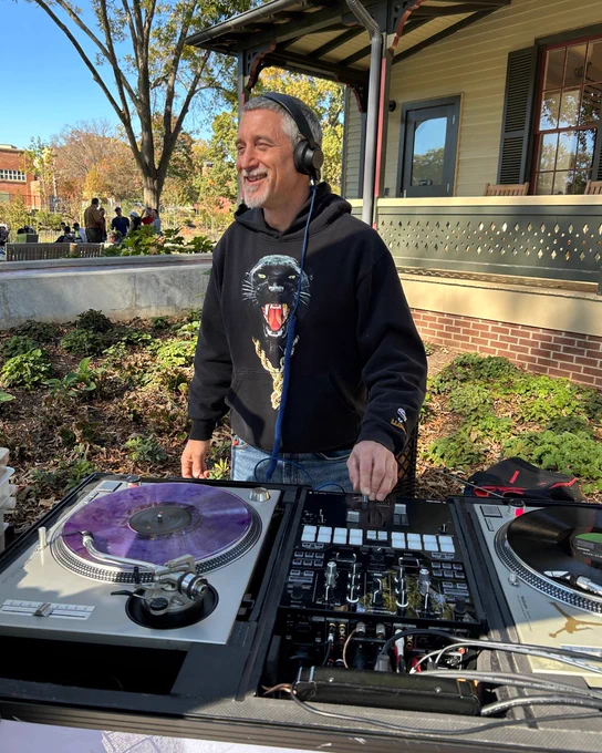 DJ wearing headphones and a black hoodie with a roaring panther graphic, mixing music on turntables outdoors near a porch.