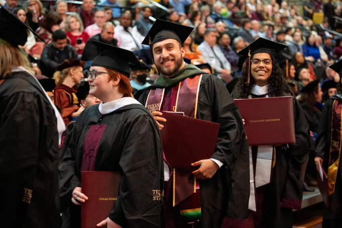 Graduates in caps and gowns holding diplomas during a university commencement ceremony with a seated audience in the background.