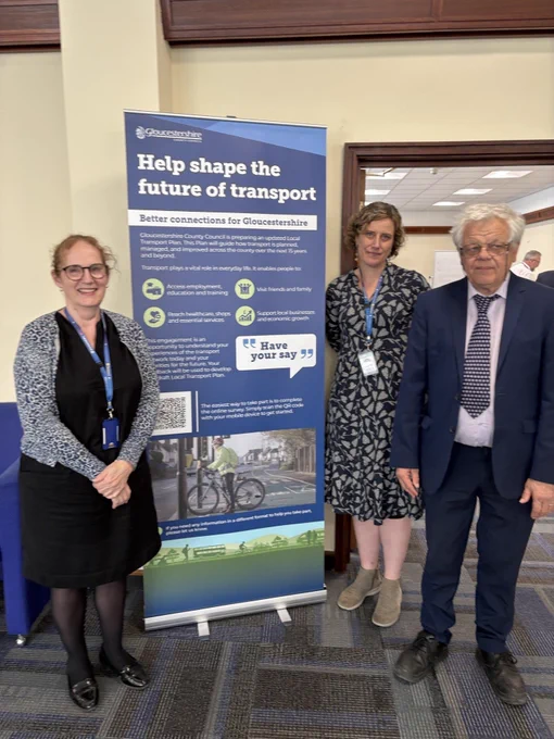 Three council representatives standing by a banner about shaping the future of transport in Gloucestershire, inviting public input.