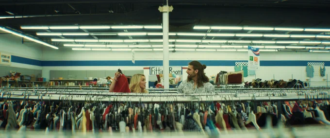 At a thrift store, a blonde woman holds up a red dress, standing near a long-haired man amidst densely packed clothing racks. 'Fitting Room' sign visible in the background, under fluorescent lighting.