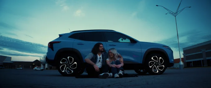 A man with long hair and a woman embrace while sitting on the ground next to a light blue car in a parking lot. The sky is blue with some clouds. Tall light pole in the background.