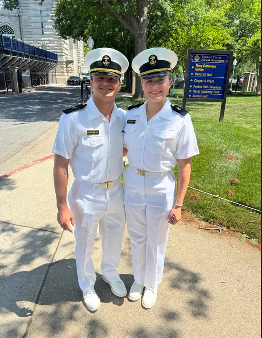 Two individuals dressed in crisp white naval uniforms stand side by side on a sidewalk. They are smiling and wearing caps. In the background, there's a building and a sign for the United States Naval Academy with a list of nearby locations.