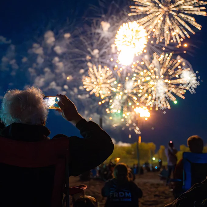 An elderly person captures bright fireworks exploding in the night sky during an outdoor celebration.