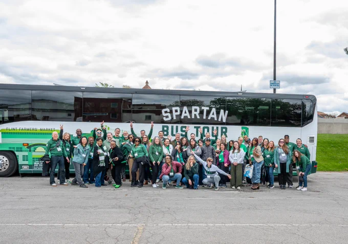Group photo of people in front of \"SPARTAN BUS TOUR\" bus. People are wearing green. Group has diverse ages and ethnicities, with various poses.