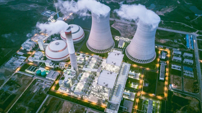 Aerial view of a large industrial facility with two cooling towers emitting steam. The complex includes several large buildings and a tall smokestack. The site is illuminated with lights and surrounded by greenery.