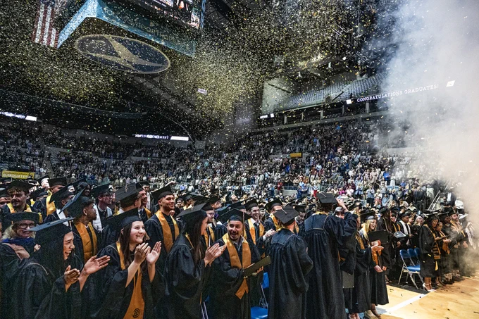 Graduates in caps and gowns celebrate at PFW commencement in the Memorial Coliseum filled with people. Confetti falls from above, and a light haze adds to the festive atmosphere.