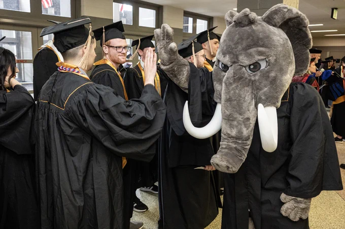 A graduate in cap and gown high fives the university mascot Don the Mastodon.