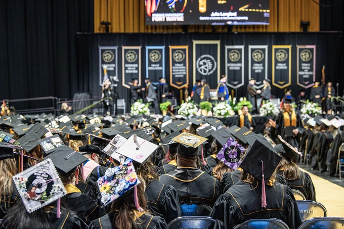 Commencement of Purdue University Fort Wayne with a large group of graduates in caps and gowns seated facing a stage. The stage features university banners and faculty members in academic regalia and many graduates' caps are festively decorated.