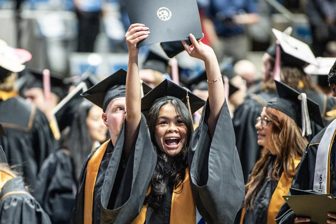 A graduate in a cap and gown joyfully raises a diploma above their head amidst a crowd of fellow graduates at PFW commencement.