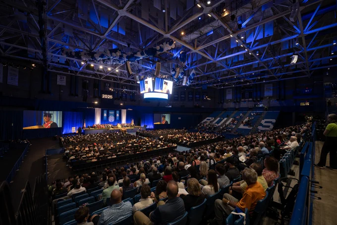 An indoor graduation ceremony. Audience members fill rows of seats. Above the stage, large screens show the speaker. The stage is set with a blue backdrop. 