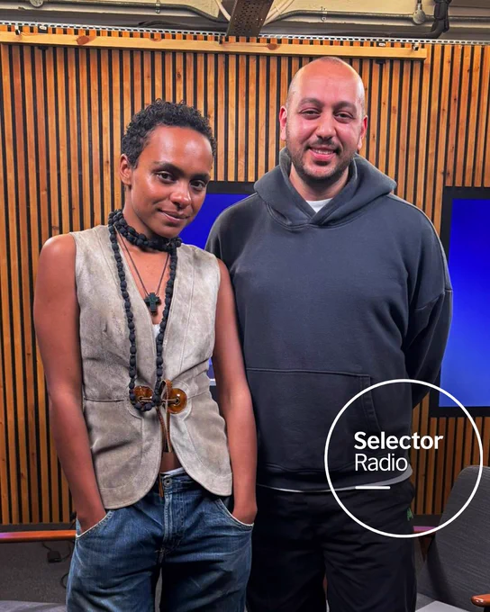 Presenter Mo Ayoub and musician Crystal Murray pose together in a wood-paneled radio studio beside two blue-lit screens and gray chairs. The Selector Radio logo appears in a white circle in the bottom right corner.