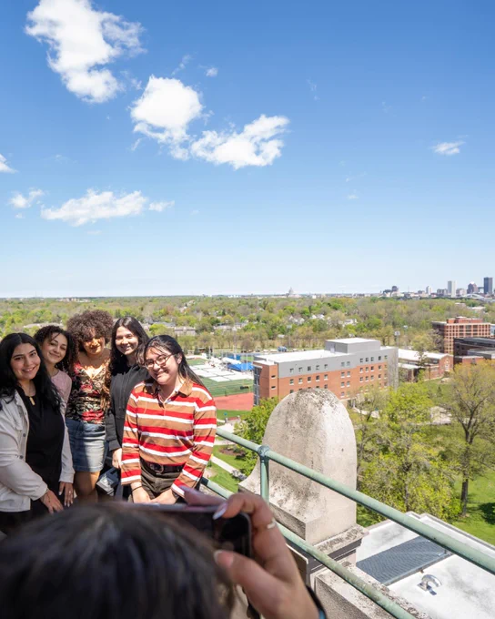 A group of students pose while another takes a photo from the top of Rush Rhees Library, with the city skyline in the background. 