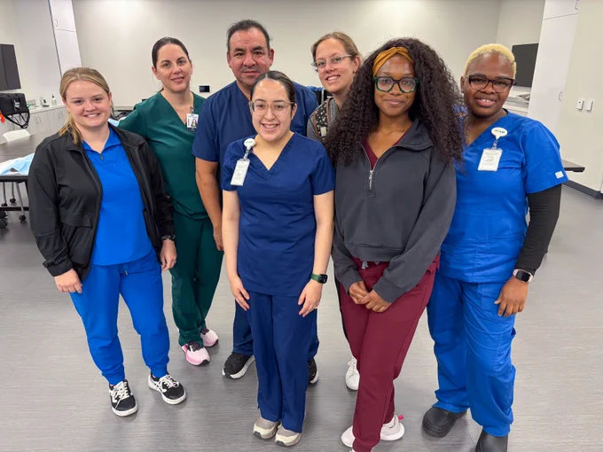 A group of seven healthcare professionals in scrubs and casual clothing pose together in a well-lit room. Each person is wearing a name badge, indicating a professional setting. The room contains medical equipment in the background.