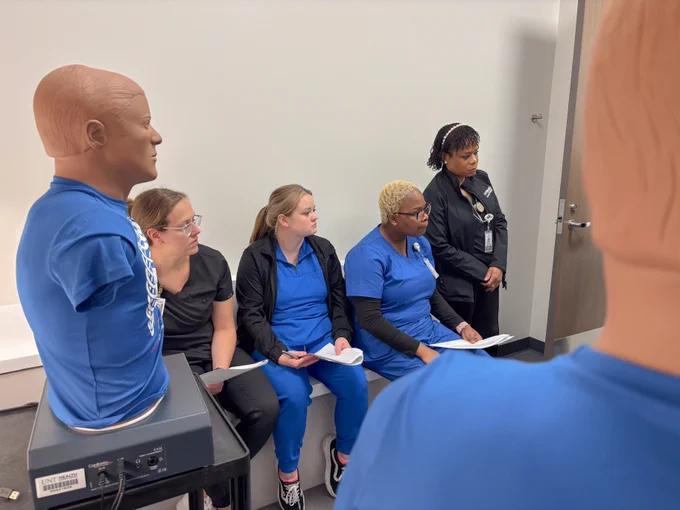 A group of individuals, some wearing blue uniforms, are seated and appear to be listening attentively during a training session. Mannequin torsos are visible in the foreground and background. A person stands near a door, possibly instructing or observing.