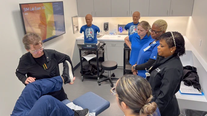 A group of people in a medical training room observe a person demonstrating a procedure on a leg. Medical equipment and training dummies are present. A monitor displays "SIM Lab Exam 1B."