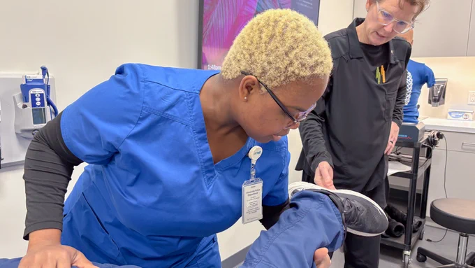 Two individuals are present in a medical setting. One person in blue scrubs is examining the leg of another person sitting while another individual in black clothing observes. Medical equipment and a poster are visible in the background.