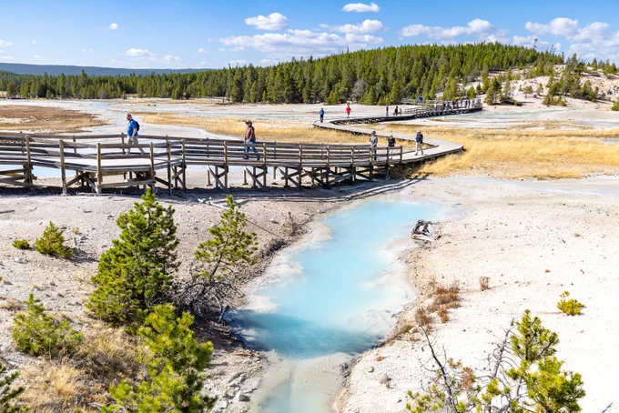 People walking on a wooden boardwalk through a hydrothermal area on a sunny summer day.
