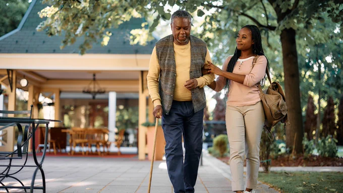 A younger person on the right is helping an older person on the left walk outside, the latter holding a cane.