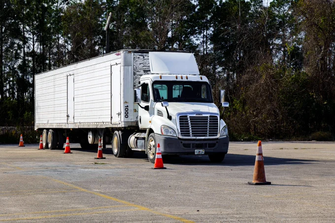 White semi-truck with a large trailer parked in an open lot surrounded by orange traffic cones and trees in the background.