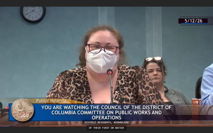 Screenshot from a public hearing at the DC Council featuring a woman with brown hair wearing glasses, an N95 face mask, and a leopard print shirt. Overlay text notes the date of 5/12/26 and reads "Public Hearing. You Are Watching the Council of the District of Columbia Committee on Public Works and Operations."