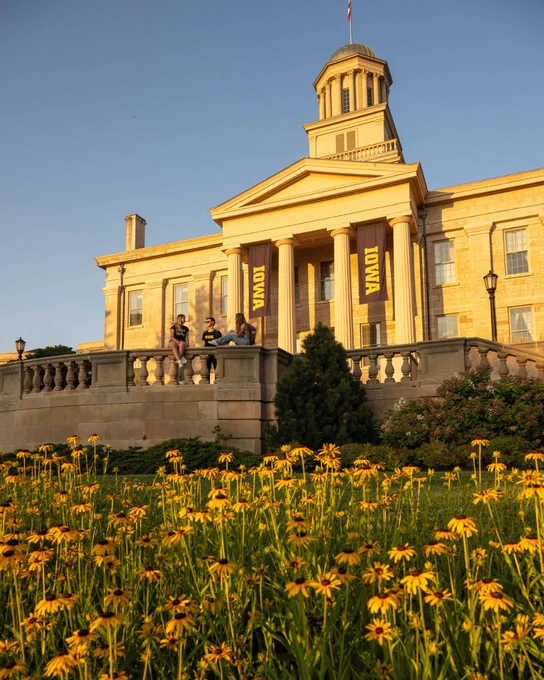 Old Capitol at the University of Iowa glows in golden evening light as students sit along the front steps and terrace. Yellow flowers bloom in the foreground, and black-and-gold Iowa banners hang between the building’s columns.