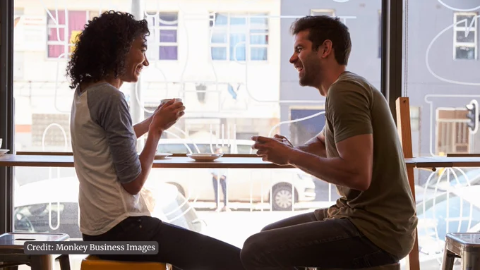 Two people sitting by a large window in a cafe, engaged in a conversation while holding coffee cups.