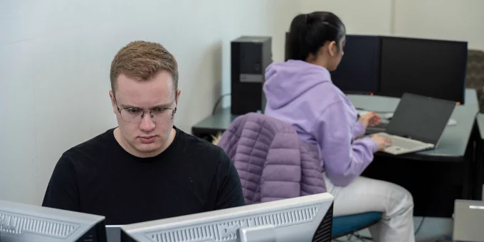 Two individuals working at computer stations. One person is focused on a monitor, while the other sits at a nearby desk with a laptop. Both are wearing casual clothing, and a purple jacket is draped over a chair.