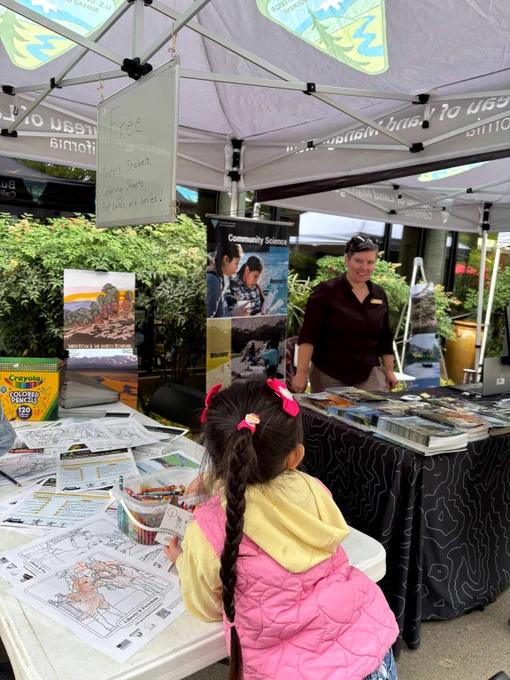 Young girl with braided hair and pink jacket coloring at a table filled with activity sheets under a canopy at an outdoor educational booth.