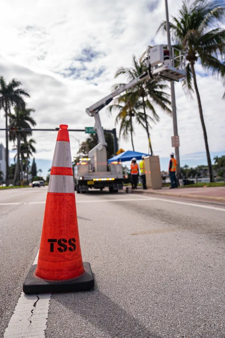 Foreground shows a \"TSS\"-labeled orange traffic cone on an asphalt road. Background shows utility workers near a truck with a raised bucket and palm trees against a cloudy sky.