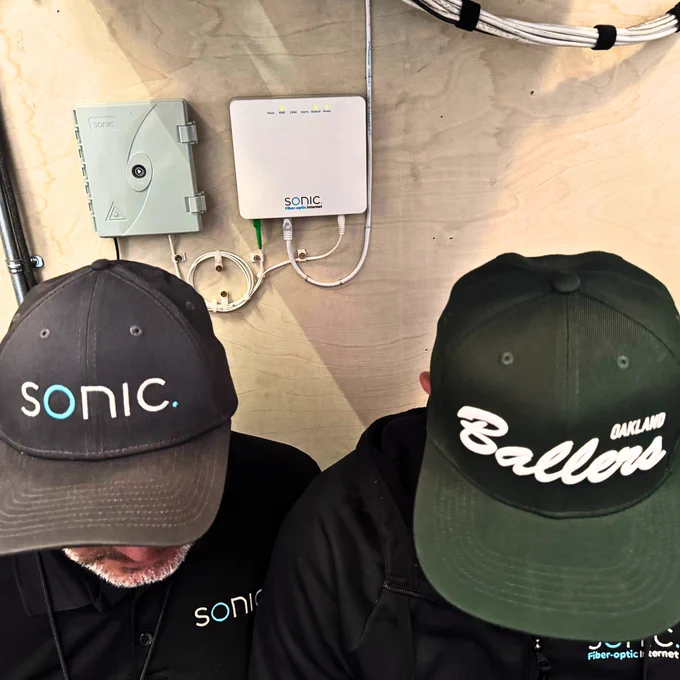 Two people are sitting on the ground indoors with their heads down. One is wearing a Sonic cap, and the other is wearing an Oakland Ballers baseball cap. Mounted on the wall behind them is a transition box and a Sonic Fiber-optic Internet ONT.  