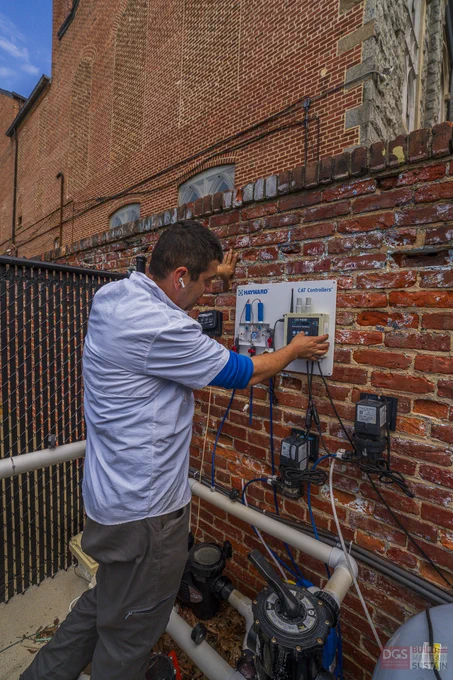 Technician adjusting a control panel mounted on a brick wall outdoors near water pipes and equipment.