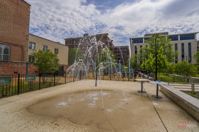 Water jets spray from a spray park in area surrounded by buildings and greenery under a partly cloudy sky.