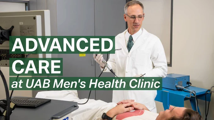 Medical exam room scene showing a clinician in a white UAB Medicine coat holding a diagnostic device with a cord while standing beside a patient lying on an exam table. The patient rests with hands folded on their abdomen as medical equipment and a computer are visible nearby. Overlaid green text reads: “ADVANCED CARE at UAB Men’s Health Clinic.”