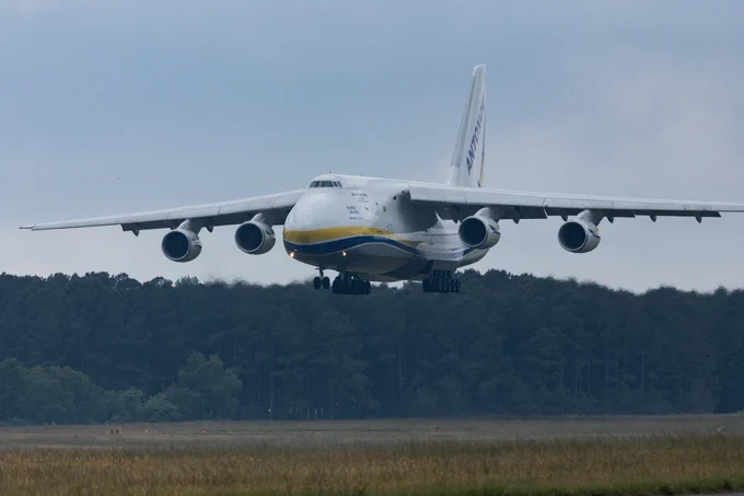The Antonov aircraft in midair before landing on the runway at Wallops. The plane is white with a yellow and blue stripe. The words \"ANTONOV\" are on the tail fin. A line of dark trees is in the background with light yellow-ish grass in the foreground. (Credit: NASA/Jamie Adkins)