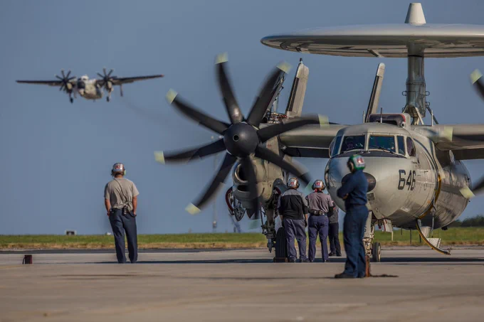 A military aircraft on a tarmac with multiple crew members around. A second military plane is seen in the distance flying in the sky. (Photo Credit: NASA/Patrick Black)