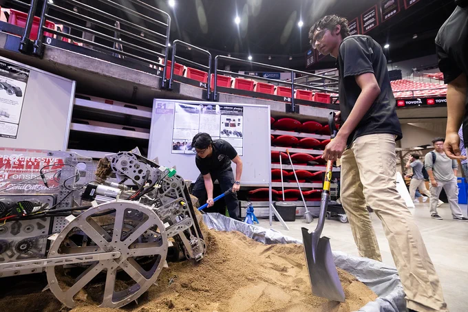A person operates a large, wheeled robot in a sandy area inside a sports arena. Another person stands nearby holding a shovel. In the background, there are rows of red seats and posters displayed on boards.