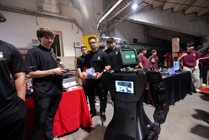 A group of people in a technology exhibition space interact with a humanoid robot. One person holds a game controller, while another holds a colorful object. The robot has a screen displaying camera feeds. 