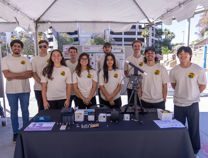 A group of individuals stand behind a table displaying various small technology devices and components under a white tent. They are all wearing light-colored shirts with a logo. 