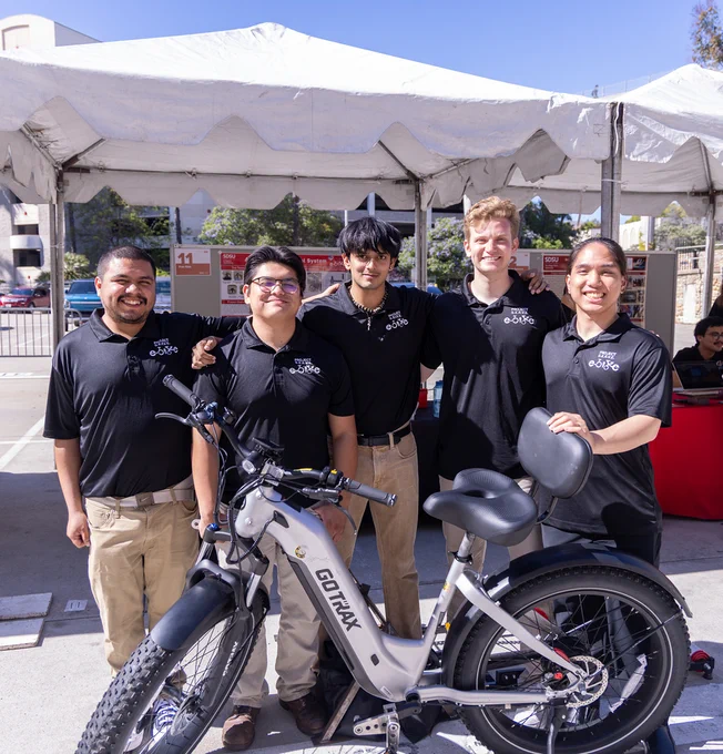 A group of five people are standing together under a white tent, smiling at the camera. They are wearing matching black polo shirts with a logo. In front of them is a Gotrax electric bike. The setting appears to be outdoors with tables in the background.